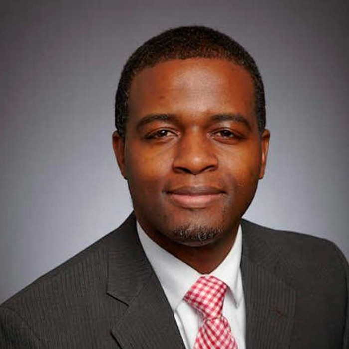 A portrait of Mark McKoy, a black man wearing a dark grey suit, white shirt, and red and white checkered tie.