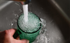 A hand holding and filling a green-tinted glass mug with water from a running faucet.