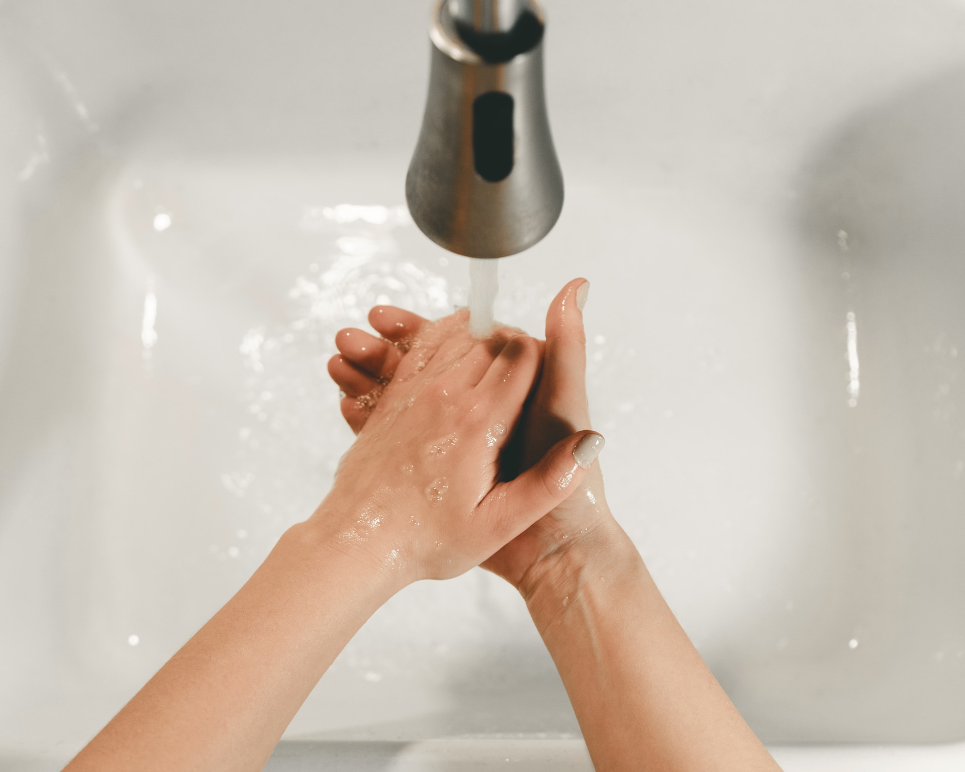 Hands being washed over a white sink with clear water running out of a metal faucet.