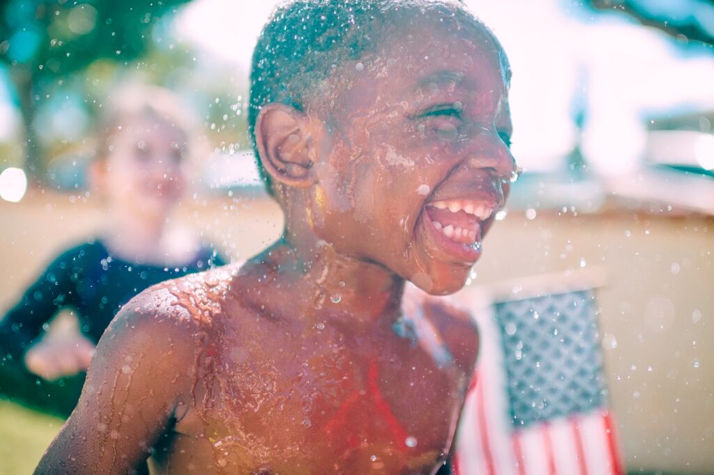A young black boy laughing and playing in a sprinkler, with a another child holding an American flag in the background.
