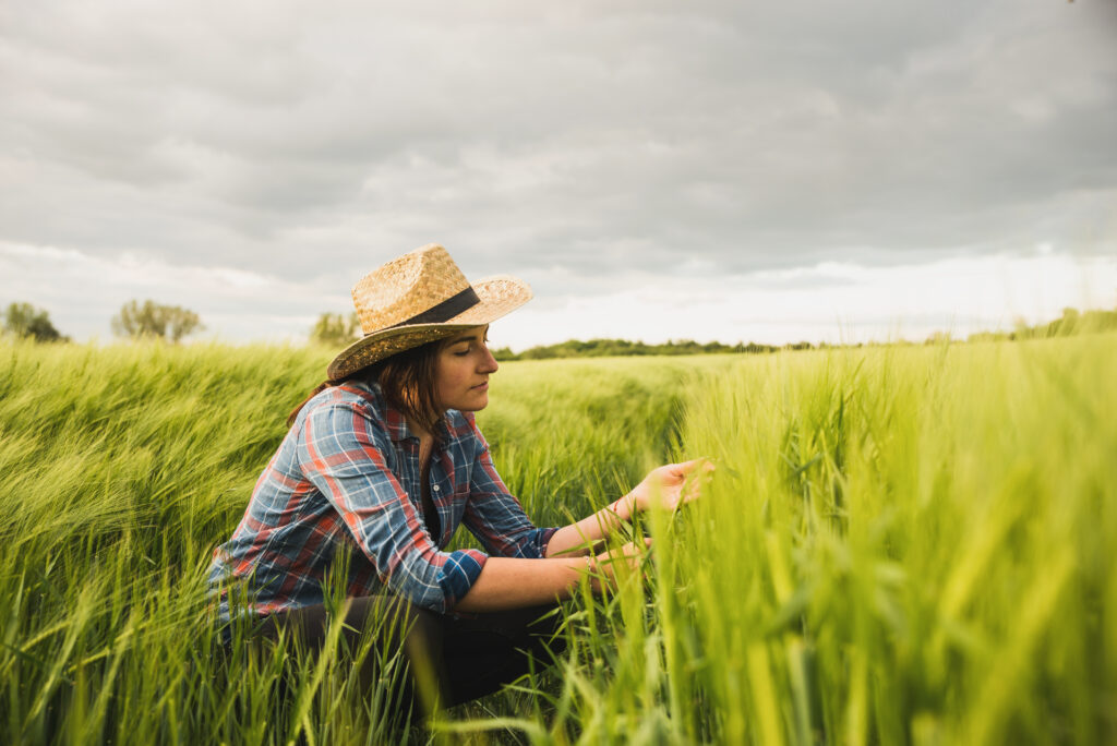 A person with long brown hair wearing a woven cowboy hat and plaid shirt, crouching in a field of tall grass.