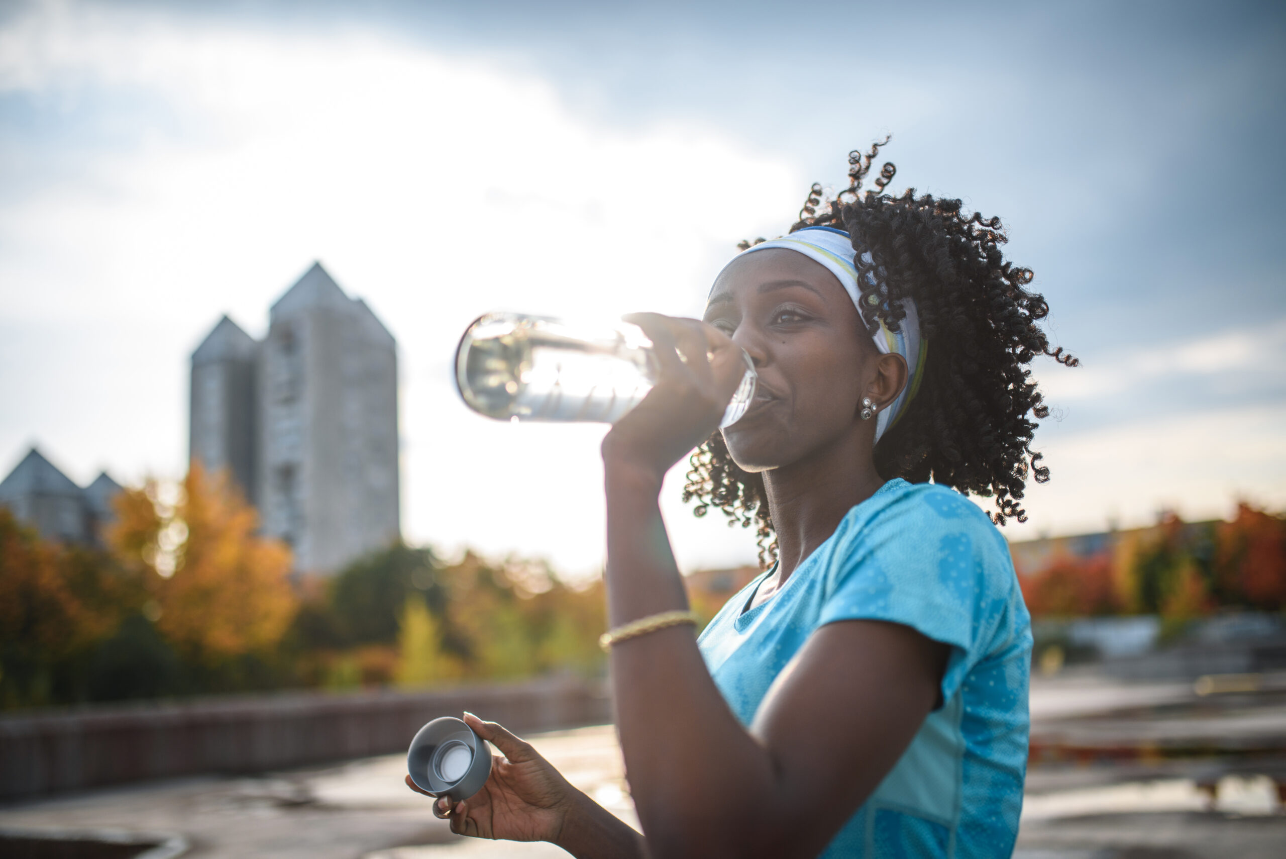 A black woman with curly natural hair, wearing a headband and blue shirt, standing outside and drinking water from a reusable bottle.