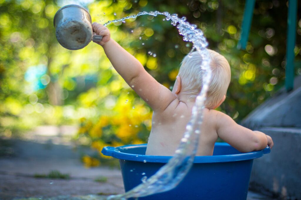 Photo of a baby sitting in a blue bucket, waving a metal cup with water spraying out behind them.