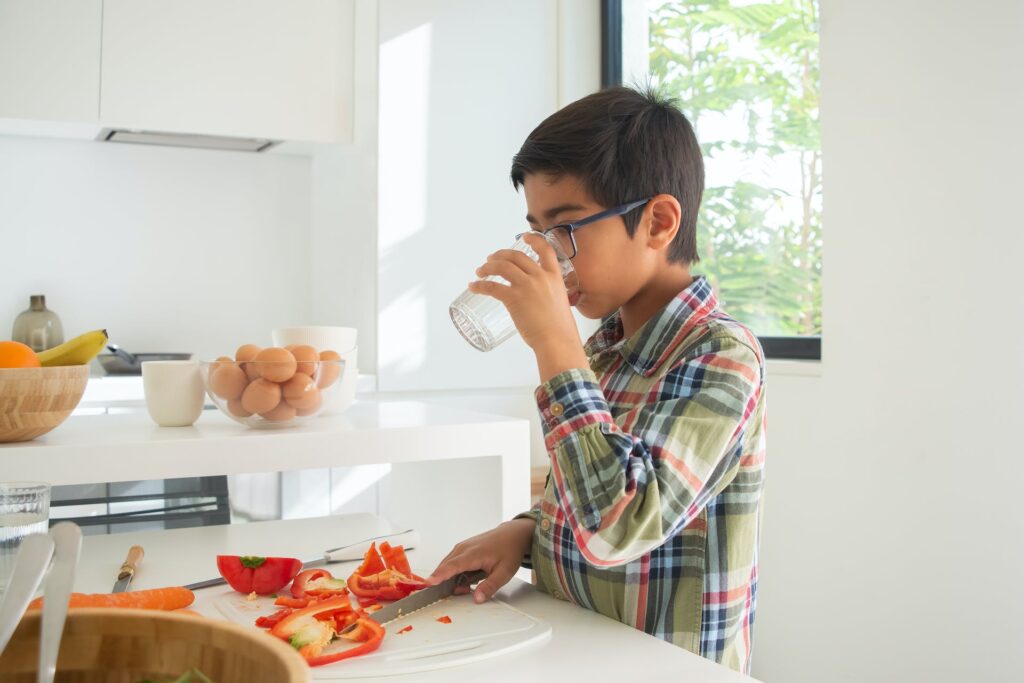 A young boy drinking water from a glass and slicing red peppers.